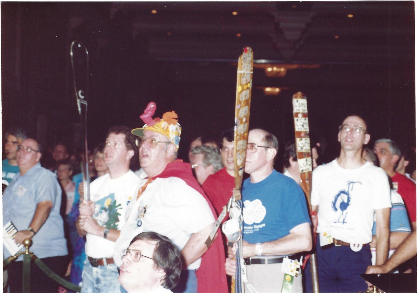in the foreground, one white man in glasses wearing a strange hate with a cape holding a clear paddle and another man holding a wood canoe paddle with stickers in a blue t-shirt and balding hairline