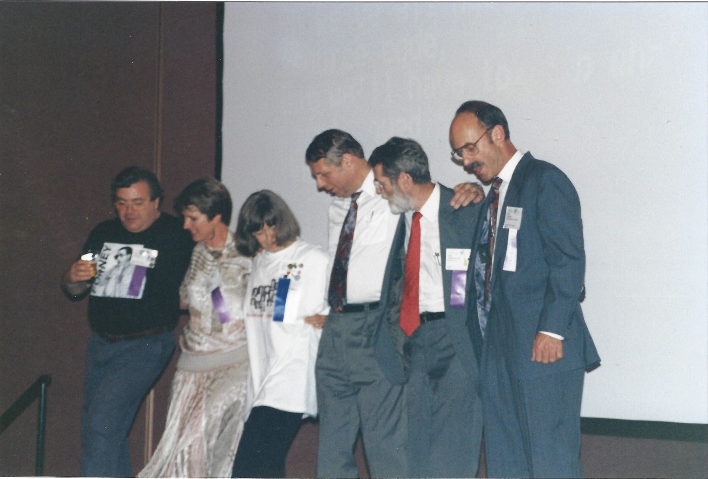 six past presidents of SHARE dancing in chorus line: first is a man with dark short hair in a t-shirt and slacks, one woman with short hair in a dress, a second woman with shoulder length hair in a t-shirt and slacks, three men in suits