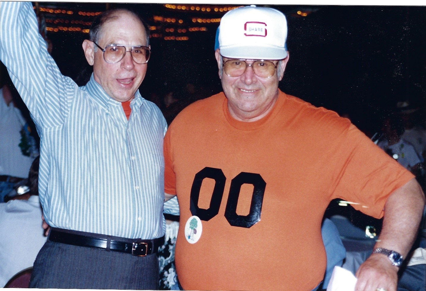 bald white man in blue and white striped dress shirt with glasses raising his hand and white man with glasses in white baseball cap and orange t-shirt grinning